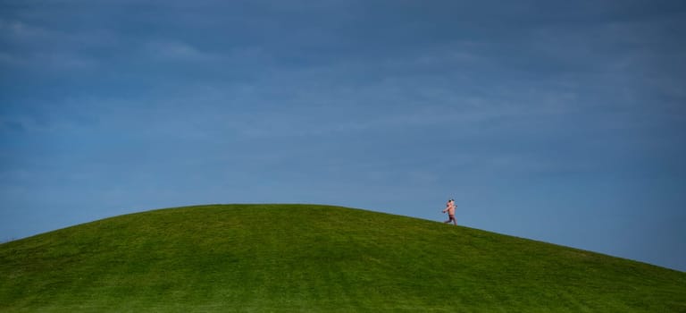 A person strolls along a vibrant green hilltop, surrounded by an expansive blue sky, experiencing tranquility and the beauty of nature in bright daylight.