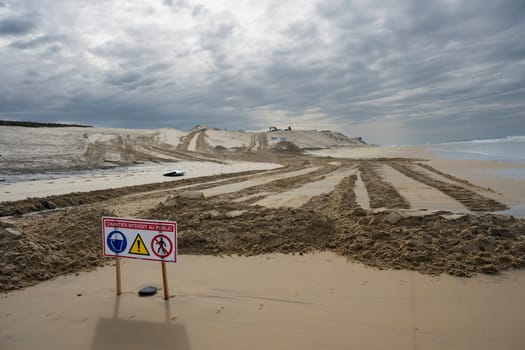 Signs indicate restricted access due to construction efforts aimed at managing coastal erosion while heavy machinery operates on the sand at Biscarosse beach under a cloudy sky.