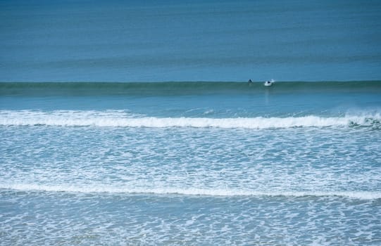Two surfers navigate the rolling waves at Biscarrosse Beach during a serene day. The calm waters reflect the azure sky, creating a perfect surfing environment.