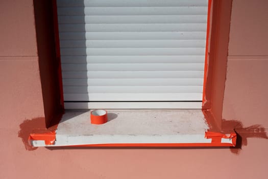 A window with white shutters is being painted with bright orange trim, showcasing fresh paint on the ledge and masking tape neatly applied around the edges for a clean finish.