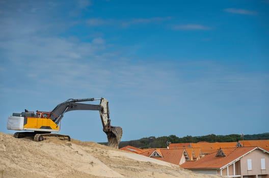 An excavator moves sand along the Biscarosse coastline under a clear blue sky. Nearby houses with orange rooftops are visible, reflecting a vibrant coastal atmosphere.