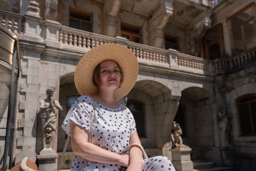 A woman wearing a straw hat and a white polka dot dress sits on a stone bench in front of a building