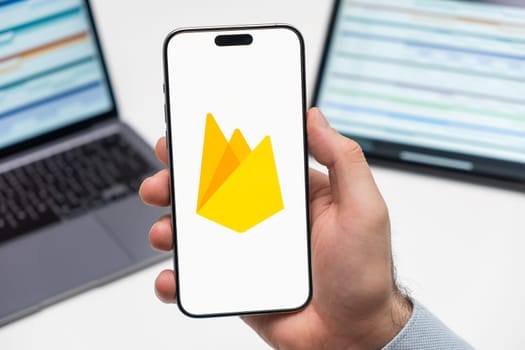 PRAGUE, CZECHIA - JULY, 2024: Firebase logo on the screen of smartphone in mans hand and laptop on the background