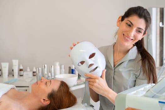 Happy female cosmetologist holds modern hydrafacial mask next to her client in salon, preparing for rejuvenating skincare treatment while creating calm and professional atmosphere