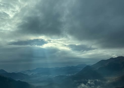A dramatic sky with thick clouds allowing beams of sunlight to stream through, illuminating the misty mountain landscape below.