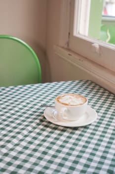 A steaming cup of coffee sits on a table with checkered cloth near a window.