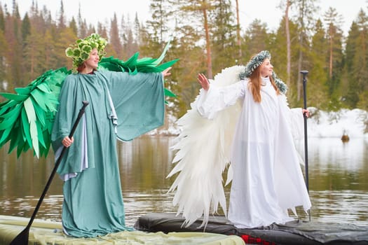 Two girls dressed in white and green dresses stand on a serene lake, embodying the harmonious meeting of winter and spring in a magical moment.