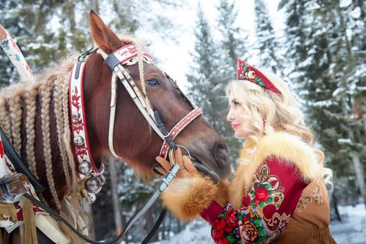 A young woman in traditional Russian clothing interacts with a horse in a winter landscape during Maslenitsa.