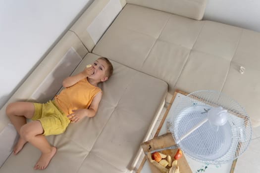 Child relaxing on sofa eating banana near fan and fruit in hot summer day