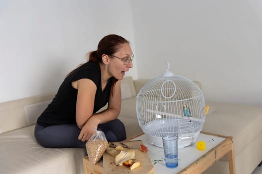 Woman preparing bird food, talking to blue budgie parrot in white cage, sitting on sofa in living room