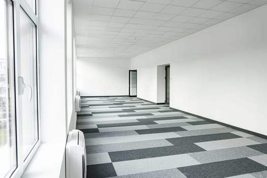 Long hallway with a checkered floor and windows on one side, allowing natural light to illuminate the space. The corridor stretches into the distance, creating a sense of depth and perspective.