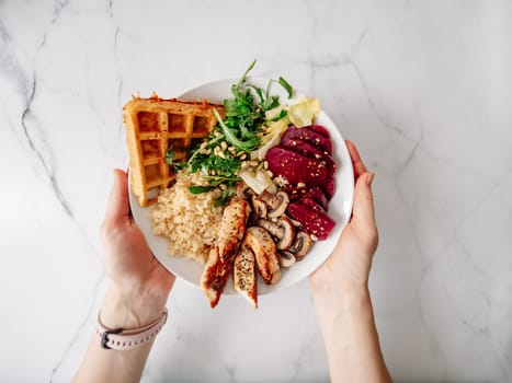 Top view of balanced gourmet dish in female hands over white marble tabletop. Bowl with chicken, mushrooms, beetroot, quinoa, green rucola, decorated pine nuts and carrot waffle. Healthy Food concept