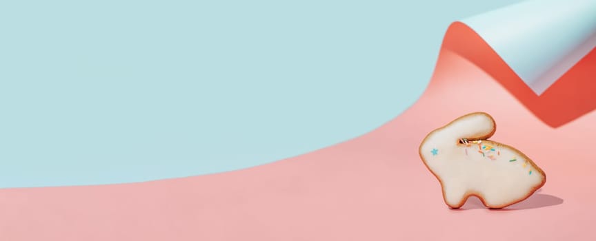Easter bunny cookie on pink and blue curved paper pastel background with shadows. Happy Easter Day concept. Minimal Easter concept. Copy space for text. Easter cooking baking background.