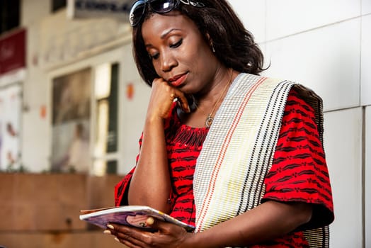 Young woman on work break, dressed in a red-black outfit, sitting against a column outside the office building, looking down, reading a notebook with her hand on her cheek.