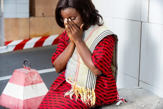 Young woman in red outfit sitting outside against office wall, looking thoughtful and frustrated, her hands on her face meditating or crying with her eyes closed on the street