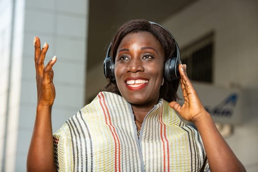 beautiful african adult woman dressed in red loincloth with a scarf around her neck is happy while having fun listening to music with headphones in the street