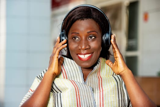 beautiful african adult woman dressed in red loincloth with a scarf around her neck is happy while having fun listening to music with headphones in the street