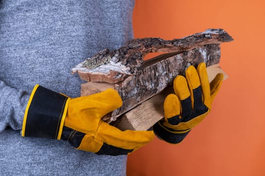 A person wearing yellow work gloves holds pieces of firewood against an orange background, emphasizing safety and outdoor work.