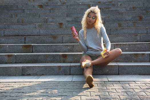 Young woman sitting on stone steps, holding a soda can and snack, enjoying a sunny day in a park setting.