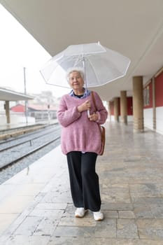 Elderly woman waiting for train arrival holding a transparent umbrella at rainy train station platform