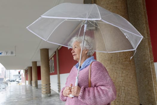 Senior woman holding a transparent umbrella walking near a building in a rainy city