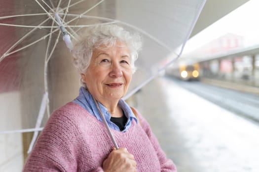 Senior woman smiling while holding a transparent umbrella at a train station on a rainy day