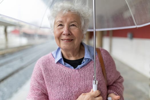 Happy elderly woman holding umbrella at train station platform on rainy day