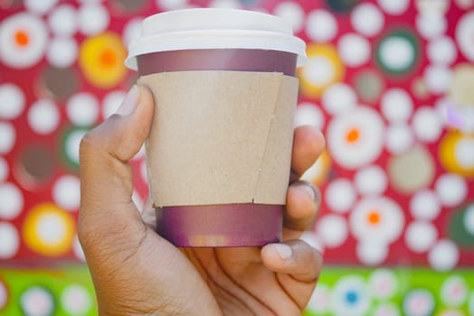 A hand grips a takeaway coffee cup against a bright, patterned backdrop.