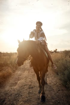 beautiful equestrian in national Ukrainian costume posing on horse. Charming girl up on brown horse in the field in the rays of the setting sun. concept of women riding horses