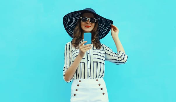 Summer portrait of beautiful happy young woman with mobile phone looking at smartphone wearing a straw tourist hat on blue studio background