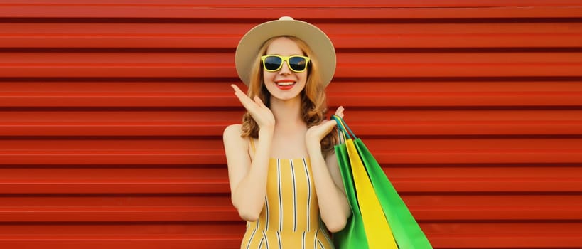 Fashion portrait, surprised happy beautiful young woman smiles posing with shopping bags in summer straw hat, looking at camera standing on red wall background