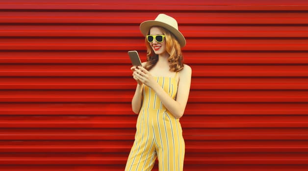 Beautiful happy young woman smiles with mobile phone looking at smartphone wearing a summer straw tourist hat on red wall background
