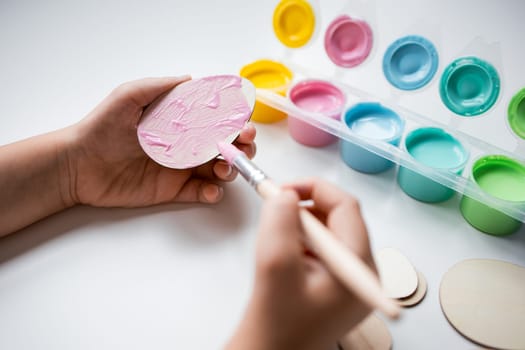 Child painting wooden egg shape with pastel colors using paintbrush on white table.