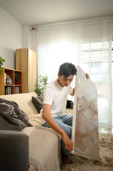 Young man removes plastic wrapping from new wooden tabletop prepares to assemble new furniture in his home.