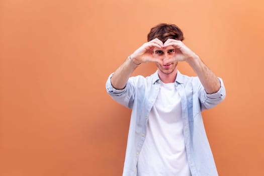 young man standing in front of orange wall doing a heart shape with hands while looking at camera, he is wearing a blue shirt and white t-shirt, concept of love, copyspace for text
