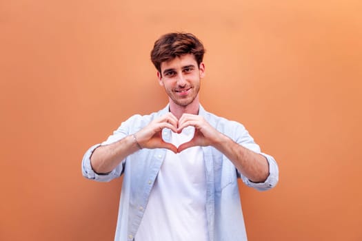 young man standing in front of orange wall doing a heart shape with hands while looking at camera, he is wearing a blue shirt and white t-shirt, concept of love, copyspace for text