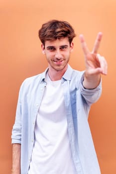 young man standing in front of orange wall holding up two fingers in victory sign and smiling looking at camera, concept of peace, love and positivity