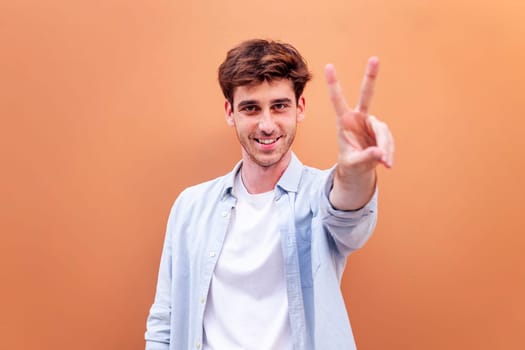 young man standing in front of orange wall holding up two fingers in victory sign and smiling looking at camera, concept of peace, love and positivity, copyspace for text