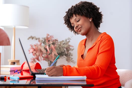 African American student studying at home, writing in notebook while using laptop. Modern workspace with stylish decor, lamp, and stationery. Education, learning, productivity, and concentration.