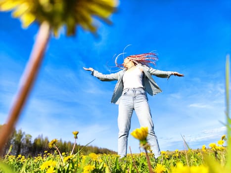 A woman with colorful hair joyfully stretches her arms wide, surrounded by blooming yellow flowers beneath a clear blue sky.