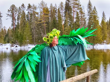 Girl wearing a green dress and wings stands by a tranquil lake, celebrating the arrival of spring and the enchanting spirit of nature.