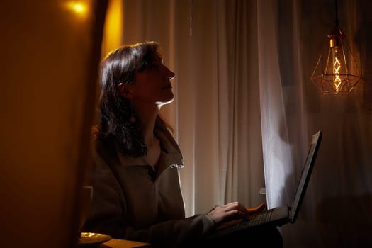 A middle-aged woman relaxes in her warm, inviting home as she engages with her laptop, appreciating the peaceful evening atmosphere.