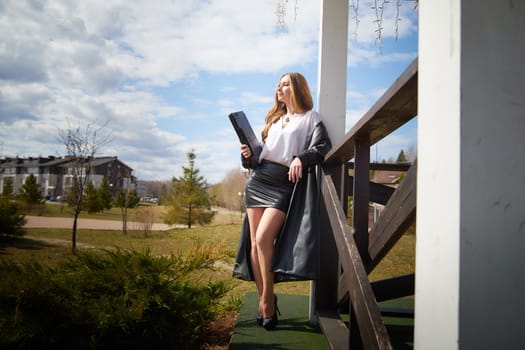 A model stands confidently on a wooden porch, dressed in a black skirt and white blouse, while holding a folder.