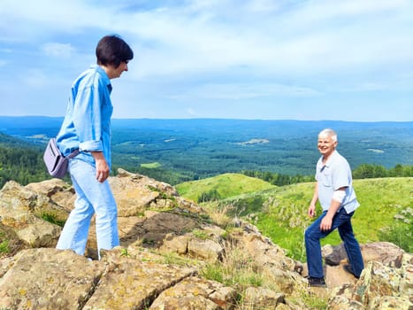 Two friends explore a rocky hilltop while surrounded by lush green valleys and a bright blue sky during summer.