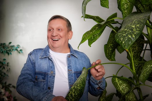A middle-aged man, smiling widely, stands in an apartment, engaging with a large indoor plant during a casual photoshoot.