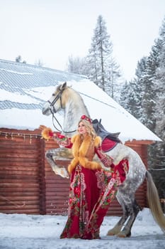 A Russian girl in colorful attire gracefully interacts with a horse in snowy scenery during Maslenitsa.