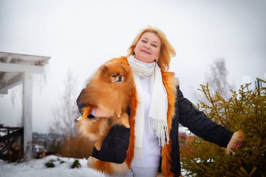 Blonde woman dressed warmly walks in the snow while holding her fluffy red Pomeranian close to her chest.