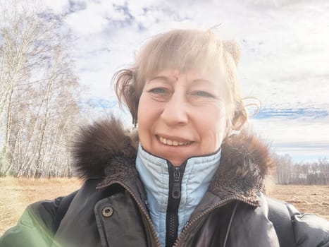 A woman smiles for a selfie while surrounded by trees under a partly cloudy sky on a clear day.
