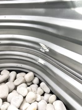 A close-up view of a corrugated metal window well with white decorative rocks at the base. The metal surface has minor scratches and wear, reflecting natural light. This functional design is commonly used for basement windows to allow light and ventilation while preventing soil erosion and water buildup.