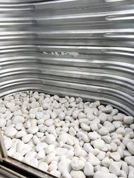 A close-up view of a corrugated metal window well with white decorative rocks at the base. The metal surface has minor scratches and wear, reflecting natural light. This functional design is commonly used for basement windows to allow light and ventilation while preventing soil erosion and water buildup.